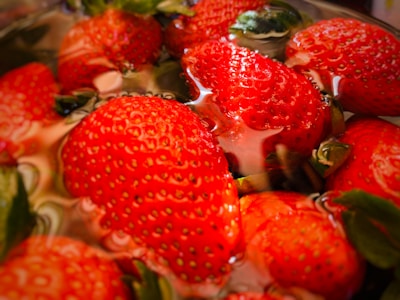 Fresh strawberries partially submerged in water, showcasing their vibrant red color and textured surface with green leaves still attached. The water creates a reflective, glossy effect on the strawberries.