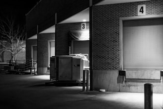 A nighttime scene depicting an industrial loading area with numbered loading dock doors made of brick. There is a metal container or dumpster placed near the dock doors, and the area is lit by a few overhead lights. In the background, a leafless tree is visible.