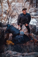 Two individuals are dressed in outdoor gear amidst a rocky landscape. One person is standing and looking upwards with a focused expression, while the other is leaning over, possibly preparing climbing equipment. Coiled ropes and a yellow bag are visible on the ground.