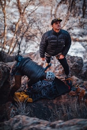 Two individuals are dressed in outdoor gear amidst a rocky landscape. One person is standing and looking upwards with a focused expression, while the other is leaning over, possibly preparing climbing equipment. Coiled ropes and a yellow bag are visible on the ground.