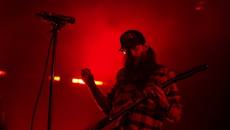 A musician wearing a checkered shirt and a cap is playing an electric guitar. The scene is bathed in red light, with a microphone stand visible beside the musician.
