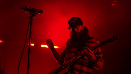 A musician wearing a checkered shirt and a cap is playing an electric guitar. The scene is bathed in red light, with a microphone stand visible beside the musician.