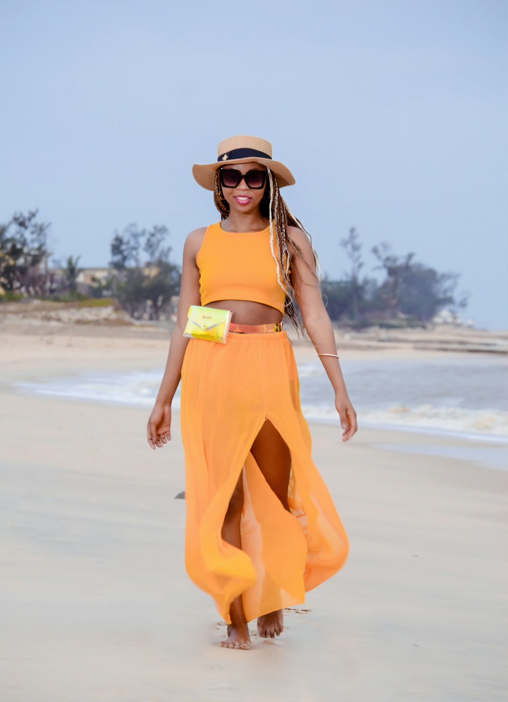 낮 동안 해변에 서 있는 갈색 태양 모자를 쓰고 노란색 스파게티 스트랩 드레스를 입은 여자