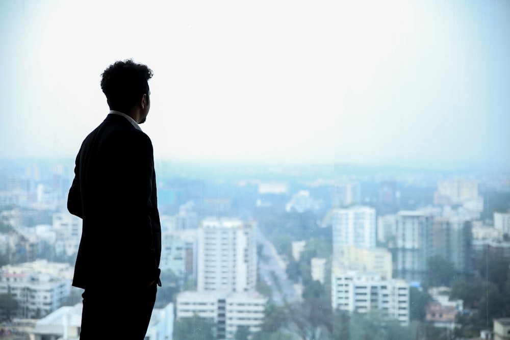 homem em terno preto em pé no topo do edifício olhando para os edifícios da cidade durante o dia