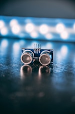 An electronic component with two round sensors is positioned on a reflective surface with a blurred background featuring bright circular lights.