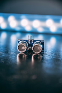 An electronic component with two round sensors is positioned on a reflective surface with a blurred background featuring bright circular lights.
