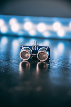 An electronic component with two round sensors is positioned on a reflective surface with a blurred background featuring bright circular lights.
