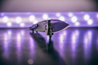 A sleek, modern knife placed on a reflective surface with a blurred background of circular lights creating a soft glow. The focus is on the detailed texture of the knife handle and its sharp blade.
