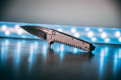 A black folding knife is placed on a reflective surface with a blurred background of blue LED lights, creating a modern and slightly tech-oriented atmosphere. The knife is angled with the blade partially open, highlighting its design and texture.
