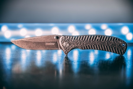 A folding knife is positioned on a reflective surface with a textured handle and a stonewashed blade. The background features a blurred pattern of circular lights, providing a contrasting backdrop.