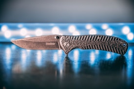 A folding knife is positioned on a reflective surface with a textured handle and a stonewashed blade. The background features a blurred pattern of circular lights, providing a contrasting backdrop.