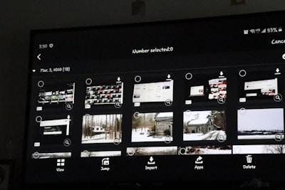A digital screen displaying a photo gallery interface with thumbnail images. The interface includes options like 'View', 'Jump', 'Import', 'Apps', and 'Delete'. The thumbnails show a mixture of screenshots and landscape photos, some of which depict snowy scenes and cabins.