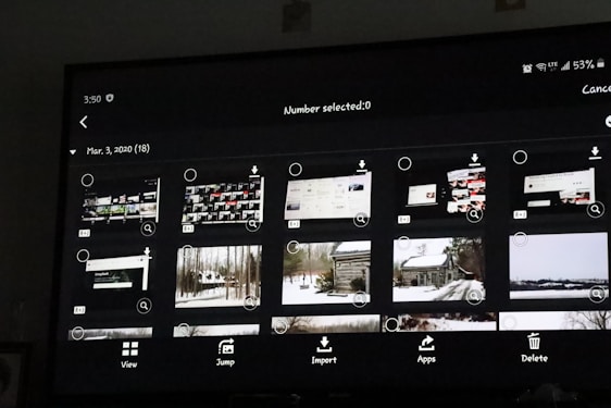 A digital screen displaying a photo gallery interface with thumbnail images. The interface includes options like 'View', 'Jump', 'Import', 'Apps', and 'Delete'. The thumbnails show a mixture of screenshots and landscape photos, some of which depict snowy scenes and cabins.
