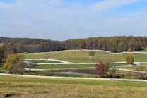 Rolling hills with a patchwork of green grass and scattered trees, bordered by a winding white wooden fence. A small pond is situated centrally, reflecting the peaceful landscape. The background features a dense forest with trees in autumnal colors, creating a serene countryside scene under a clear blue sky.