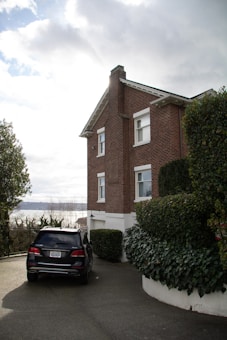 A brick two-story house with a chimney is situated on a sloped driveway surrounded by well-maintained bushes and hedges. A black SUV is parked in front of the house, and in the background, a body of water is visible under a cloudy sky.