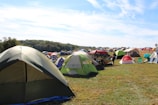A large open field filled with numerous colorful tents set up for camping. The tents vary in color and size, with green, red, and beige prominent among them. The grassy area is surrounded by a wooded landscape under a partly cloudy blue sky. There are a few folding chairs scattered around the tents, and a person is visible near one of the tents, suggesting a relaxed, recreational activity or festival.