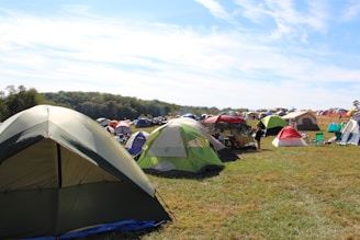 A large open field filled with numerous colorful tents set up for camping. The tents vary in color and size, with green, red, and beige prominent among them. The grassy area is surrounded by a wooded landscape under a partly cloudy blue sky. There are a few folding chairs scattered around the tents, and a person is visible near one of the tents, suggesting a relaxed, recreational activity or festival.