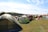 A large open field filled with numerous colorful tents set up for camping. The tents vary in color and size, with green, red, and beige prominent among them. The grassy area is surrounded by a wooded landscape under a partly cloudy blue sky. There are a few folding chairs scattered around the tents, and a person is visible near one of the tents, suggesting a relaxed, recreational activity or festival.