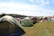 A large open field filled with numerous colorful tents set up for camping. The tents vary in color and size, with green, red, and beige prominent among them. The grassy area is surrounded by a wooded landscape under a partly cloudy blue sky. There are a few folding chairs scattered around the tents, and a person is visible near one of the tents, suggesting a relaxed, recreational activity or festival.