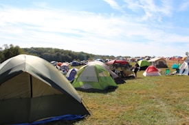 A large open field filled with numerous colorful tents set up for camping. The tents vary in color and size, with green, red, and beige prominent among them. The grassy area is surrounded by a wooded landscape under a partly cloudy blue sky. There are a few folding chairs scattered around the tents, and a person is visible near one of the tents, suggesting a relaxed, recreational activity or festival.