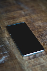 A smartphone with a black screen is placed on a wooden surface. The phone lies horizontally with a slight angle, highlighting its sleek design and glossy finish.