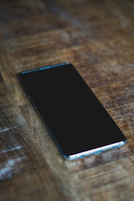 A smartphone with a black screen is placed on a wooden surface. The phone lies horizontally with a slight angle, highlighting its sleek design and glossy finish.