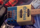 A close-up of a unique bass guitar made from a recycled metal container, labeled with 'Surfwax' and additional text. The guitar has four strings and visible tuning pegs, with a microphone cord plugged into it. The background includes glimpses of a person wearing a plaid shirt.