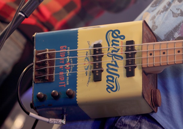 A close-up of a unique bass guitar made from a recycled metal container, labeled with 'Surfwax' and additional text. The guitar has four strings and visible tuning pegs, with a microphone cord plugged into it. The background includes glimpses of a person wearing a plaid shirt.