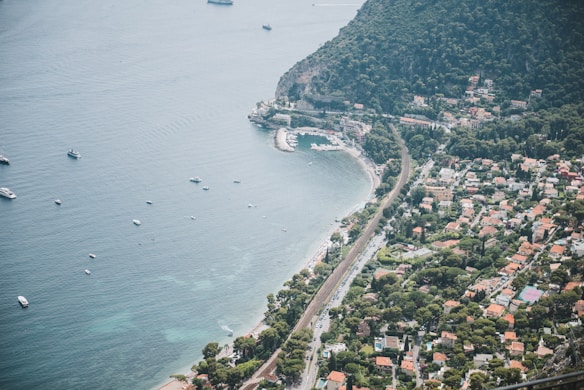 A coastal town is visible from an aerial perspective, showcasing a blend of urban development and natural landscape. The shoreline curves gently, with clear, turquoise waters dotted with small boats. The town is nestled between lush green hills and the sea, characterized by a mix of residential buildings with red-tiled roofs. A railway line runs parallel to the coastline, and there are roads with vehicles moving along them.