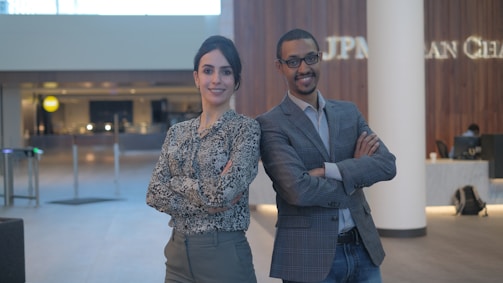 A man and a woman standing confidently with their arms crossed in a modern office lobby environment. The background features wooden wall panels and a reception area with soft lighting.