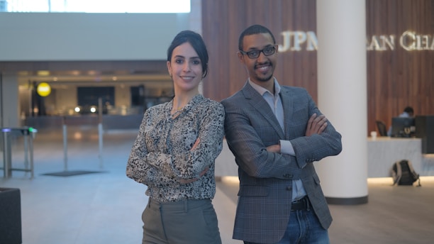 A man and a woman standing confidently with their arms crossed in a modern office lobby environment. The background features wooden wall panels and a reception area with soft lighting.