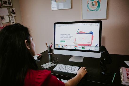 A person is sitting at a desk using a large desktop computer with a website open on the screen. The desk has a microphone, a keyboard, and other office supplies. There are some framed artworks on the wall and a bulletin board on the side displaying several photos. The mood is busy and focused, suggesting a work-from-home or creative workspace environment.