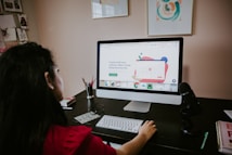 A person is sitting at a desk using a large desktop computer with a website open on the screen. The desk has a microphone, a keyboard, and other office supplies. There are some framed artworks on the wall and a bulletin board on the side displaying several photos. The mood is busy and focused, suggesting a work-from-home or creative workspace environment.