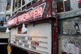 A vintage-looking theater sign with neon lights displaying 'Market Theater'. Below it, there is a board listing show times for theatre sports at 10:30, improv comedy at 8:30 PM, and improv happy hour at 7 PM. The signboard and the surrounding walls show signs of wear and age, with some peeling paint and old posters.