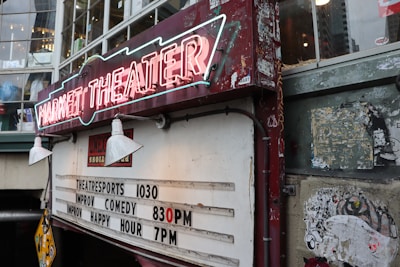 A vintage-looking theater sign with neon lights displaying 'Market Theater'. Below it, there is a board listing show times for theatre sports at 10:30, improv comedy at 8:30 PM, and improv happy hour at 7 PM. The signboard and the surrounding walls show signs of wear and age, with some peeling paint and old posters.