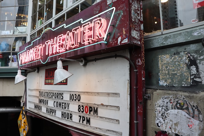 A vintage-looking theater sign with neon lights displaying 'Market Theater'. Below it, there is a board listing show times for theatre sports at 10:30, improv comedy at 8:30 PM, and improv happy hour at 7 PM. The signboard and the surrounding walls show signs of wear and age, with some peeling paint and old posters.