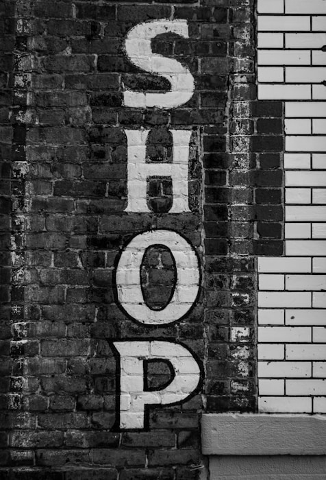 Large white block letters spelling 'SHOP' are painted vertically on a textured brick wall. The wall is a mix of old grey bricks and white tile, creating a vintage urban look.