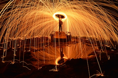 A person stands on a raised platform or structure, spinning steel wool that creates a stunning display of orange and yellow sparks in an arc around them. The reflection of the scene is visible in a pool of water below, enhancing the visual effect. The background is dark, highlighting the vibrant light display.