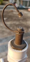A close-up of a rusty metal valve lever attached to a white plastic pipe. The metal has a rough texture and a curved handle ending in a solid piece. The background is blurred and appears to be an outdoor setting, possibly a rooftop or concrete surface.