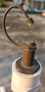 A close-up of a rusty metal valve lever attached to a white plastic pipe. The metal has a rough texture and a curved handle ending in a solid piece. The background is blurred and appears to be an outdoor setting, possibly a rooftop or concrete surface.