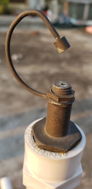 A close-up of a rusty metal valve lever attached to a white plastic pipe. The metal has a rough texture and a curved handle ending in a solid piece. The background is blurred and appears to be an outdoor setting, possibly a rooftop or concrete surface.