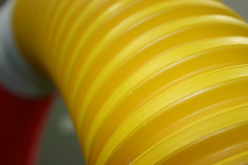 A bright yellow corrugated tube with ribbed texture. The background is slightly out of focus, featuring hints of red and white colors.