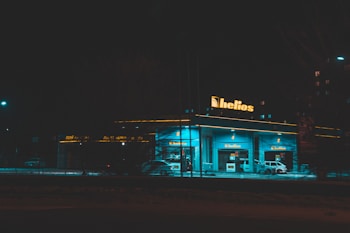 A gas station at night illuminated by vibrant blue-green lighting with the sign 'helios' prominently displayed on top. There are several fuel pumps visible and a few cars parked near them. The surrounding area is dimly lit, highlighting the glow from the station.