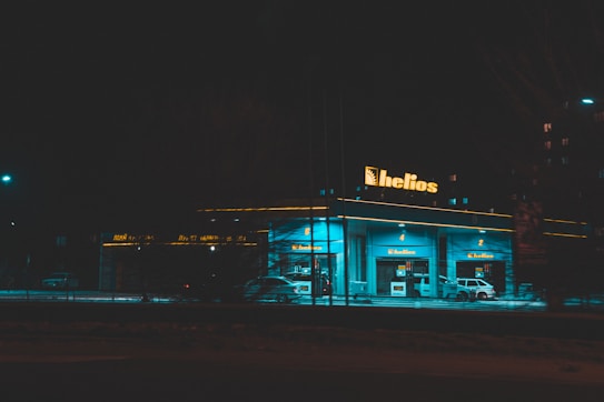 A gas station at night illuminated by vibrant blue-green lighting with the sign 'helios' prominently displayed on top. There are several fuel pumps visible and a few cars parked near them. The surrounding area is dimly lit, highlighting the glow from the station.