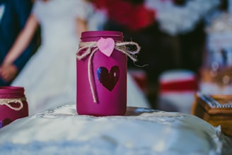 A purple jar decorated with a heart shape and tied with twine and a pink heart tag is placed on a soft, white cushion. In the blurred background, a couple appears dressed in formal clothing, possibly a bride and groom.