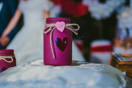 A purple jar decorated with a heart shape and tied with twine and a pink heart tag is placed on a soft, white cushion. In the blurred background, a couple appears dressed in formal clothing, possibly a bride and groom.