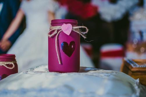 A purple jar decorated with a heart shape and tied with twine and a pink heart tag is placed on a soft, white cushion. In the blurred background, a couple appears dressed in formal clothing, possibly a bride and groom.