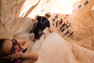 People are climbing down a steep, narrow canyon with sandy-colored rock walls. A person in the foreground looks excited or anxious with hands on cheeks, wearing sunglasses. Several individuals are visible further down the canyon path, some wearing backpacks and hats.