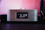 A digital clock is prominently displayed with a black and white interface, showing the time as 5:38 PM on a Tuesday, with 'Mart' mentioned below the time. The clock has a modern design with a metallic mesh exterior, placed against a pinkish background, suggesting it's on a desk or bedside table.
