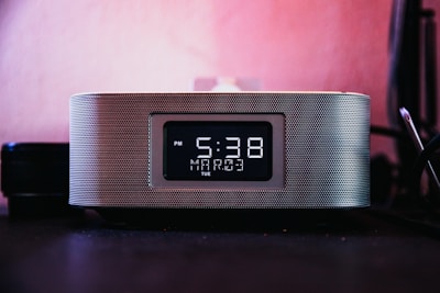 A digital clock is prominently displayed with a black and white interface, showing the time as 5:38 PM on a Tuesday, with 'Mart' mentioned below the time. The clock has a modern design with a metallic mesh exterior, placed against a pinkish background, suggesting it's on a desk or bedside table.